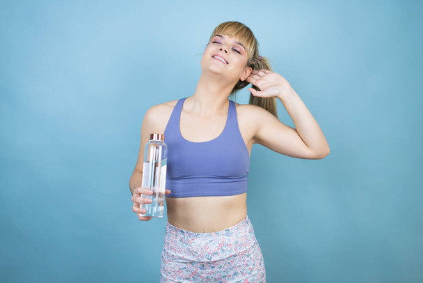 Spor kıyafetleri giymiş genç bir kadın izole edilmiş mavi arka planda bir şişe su tutuyor rahatlıyor ve esniyor, kolları ve elleri başının ve boynunun arkasında mutlu bir şekilde gülümsüyor. - Fotoğraf, Görsel
