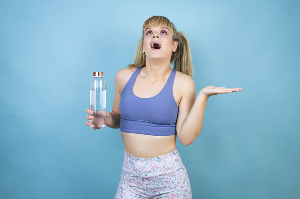 Joven hermosa mujer usando ropa deportiva sosteniendo una botella de agua sobre fondo azul aislado expresión despistada y confusa con brazos y manos levantadas - Foto, Imagen