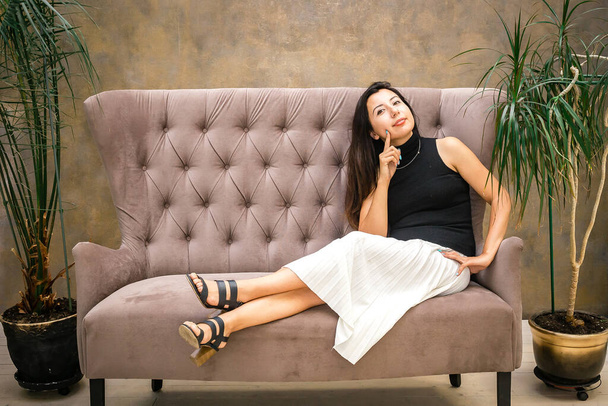 Woman with dark hair in a white skirt sits on the sofa and smiles. - Foto, Imagen