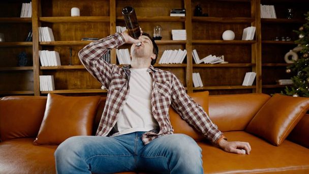 Alcohol addiction at young age. Caucasian male cannot stop drinking alcohol, drunken lonely male drinks wine from bottle. Loneliness, depression and alcoholism during Christmas and New Year holidays. - Photo, Image