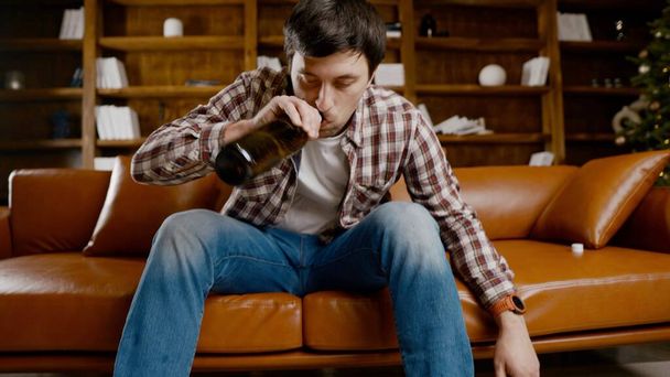 若い男が家で酒を飲んで落ち込んでいる。失業中の男性は、クリスマスツリーの近くのソファの上で直接ワインを飲み続けます。中毒、孤独、神経衰弱新年の休日. - 写真・画像