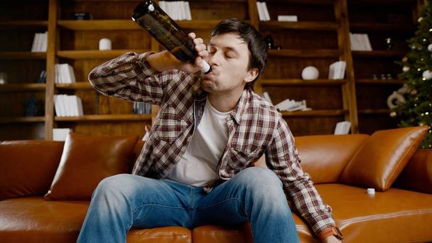 Young male alcoholic requires medical and psychological help. Man addicted alcohol and cannot stay on couch at home on Christmas and New Year Eve. Drunk guy drink wine from bottle near Christmas tree. - Photo, Image