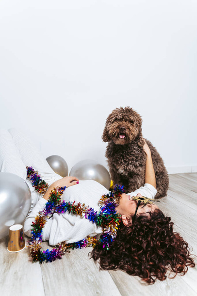 Barna nő pizsamában feküdt a földön körülvéve lufikkal ünnepli az újévet a spanyol vízi kutyájával otthon. 2021 Szilveszteri party otthon koncepció - Fotó, kép