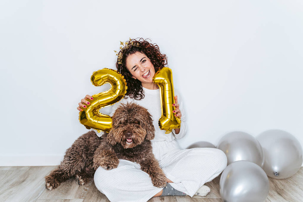 Güzel bir kadın, İspanyol su köpeğiyle pijamalarıyla yerde oturuyor ve yılbaşı gecesini evde kutlarken kameraya bakıyor. 2021 Yeni Yıl Partisi ev kavramında - Fotoğraf, Görsel