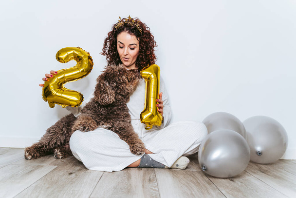 Genç bir kadın köpeğiyle yerde oturuyor ve yılbaşı partisinde altın balonlarla oynuyor. O, dikkat ederken köpeğine bakıyor. Yeni Yıl Arifesi ev kavramında - Fotoğraf, Görsel