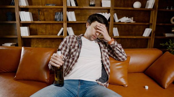 Young male alcoholic requires medical and psychological help. Man addicted alcohol and cannot stay on couch at home on Christmas and New Year Eve. Drunk guy drink wine from bottle near Christmas tree. - Photo, Image