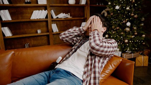Depressiver junger Mann, der zu Hause Alkohol trinkt. Arbeitslose männlich, betrunken und weiterhin trinken Weinflasche direkt auf dem Sofa in der Nähe des Weihnachtsbaums. Sucht, Einsamkeit, Nervenzusammenbruch. - Foto, Bild