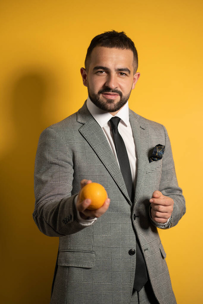 Przystojny elegancki mężczyzna w szarym garniturze w okularach wyciąga rękę oferując pomarańczę w jednej ręce stojącej odizolowany na żółtym tle. Moda męska - Zdjęcie, obraz