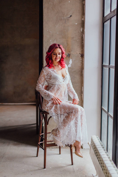Attractive woman with pink hair in white light dress posing on the bar chair near large window. Lady in gentle lace peignoir sitting in a dark room with concrete floor and walls - Foto, immagini