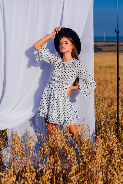 Rural Countryside Scene. Young beautiful woman with long hair dressed in white Polka-dot dress and black hat standing at golden oat field with a white curtain on the background. - Фото, изображение