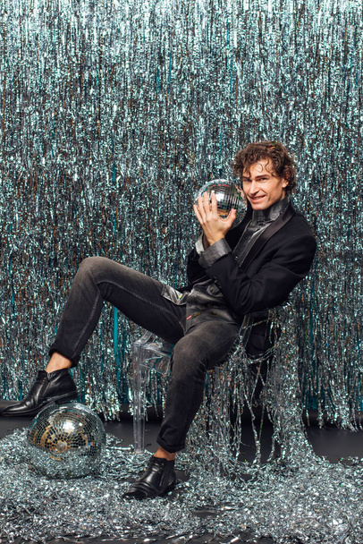Tall attractive man sitting on the chair over the sparkle background holding mirror disco ball. Party concept. - Foto, afbeelding