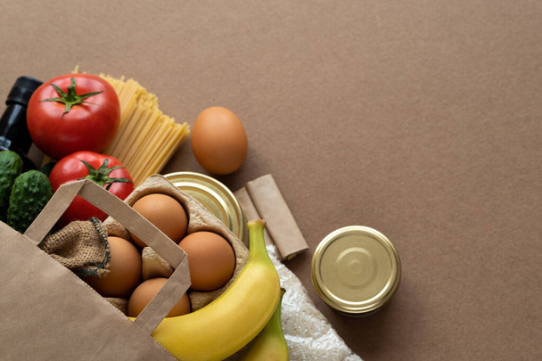 Paper bag with food supplies on neutral beige background. Pasta, rice, oil, vinegar, eggs, bananas, tomatoes, preserves, cucumbers, spices. Delivery for quarantine, donation concept. Copy space text - Photo, image