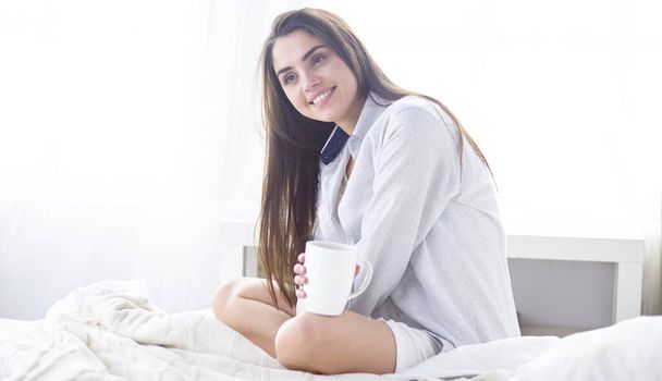 Young beautiful happy brunette woman in black silk dressing gown lying in bed, drinking morning coffee, white cup. Womens day holiday - Photo, Image