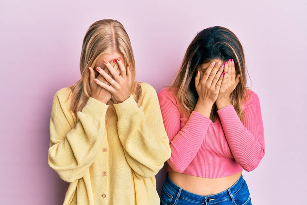 Two friends standing together over pink background with sad expression covering face with hands while crying. depression concept.  - Фото, зображення