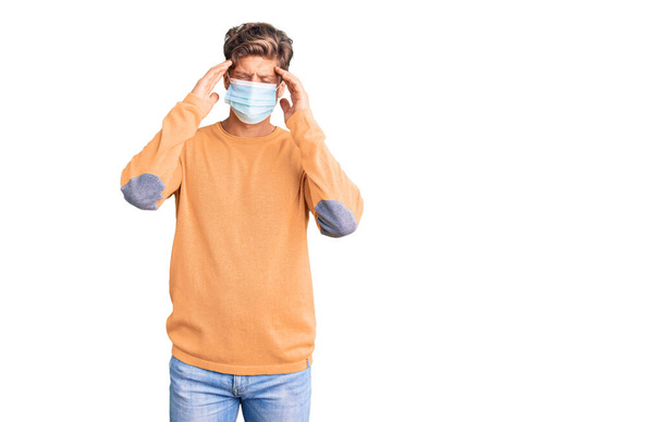 Jonge knappe man met een medisch masker die lijdt aan hoofdpijn wanhopig en gestrest omdat pijn en migraine. handen op het hoofd.  - Foto, afbeelding