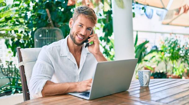 Terasta oturan yakışıklı beyaz adam evde bilgisayarla çalışıyor ve telefonla konuşuyor. - Fotoğraf, Görsel
