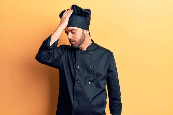 Junger hispanischer Mann in Kochuniform überrascht mit Hand auf Kopf für Fehler, erinnere dich an Fehler. Vergessenes, schlechtes Gedächtnis.  - Foto, Bild