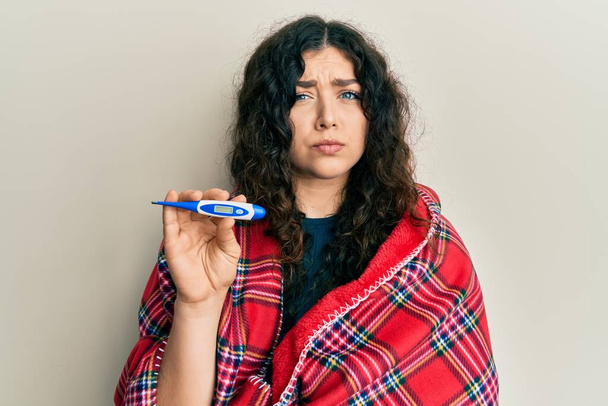 Young brunette woman with curly hair wrapped in a blanket holding thermometer skeptic and nervous, frowning upset because of problem. negative person.  - Fotó, kép