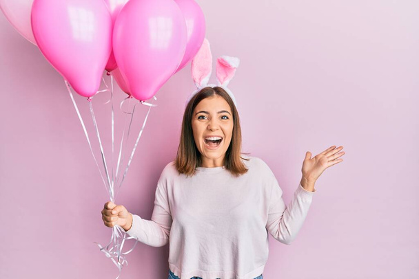 Jonge mooie vrouw draagt schattige paashaas oren en het houden van ballonnen vieren overwinning met een vrolijke glimlach en winnaar uitdrukking met opgeheven handen  - Foto, afbeelding