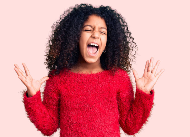 Αφροαμερικανό παιδί με σγουρά μαλλιά που φοράει ένα απλό χειμωνιάτικο πουλόβερ που γιορτάζει τρελά και τρελά την επιτυχία με τα χέρια σηκωμένα και τα κλειστά μάτια να ουρλιάζουν ενθουσιασμένα. έννοια νικητή  - Φωτογραφία, εικόνα