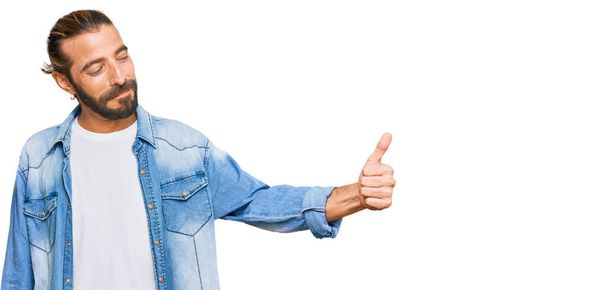Attraktiver Mann mit langen Haaren und Bart in lässiger Jeansjacke, der stolz schaut, lächelnd Daumen nach oben zur Seite streckt  - Foto, Bild
