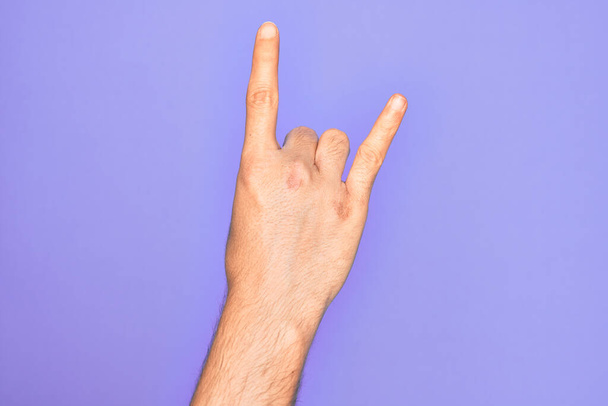 İzole edilmiş mor arka planda parmaklarını gösteren beyaz bir gencin eli. Rock 'n Roll sembolü, müstehcen boynuzlar gösteriyor. - Fotoğraf, Görsel