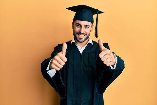 Молодой латиноамериканец в шапочке для выпускного и в свадебном халате, одобряя позитивный жест рукой, улыбается и радуется успеху. жест победителя.  - Фото, изображение