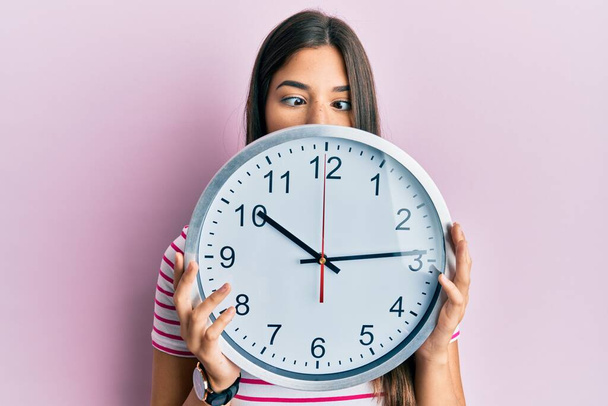 Young brunette woman holding big clock covering face making fish face with mouth and squinting eyes, crazy and comical.  - Foto, Imagen