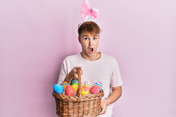 Молодий білий чоловік у милому великодні вуха кролика тримає плетений кошик з кольоровими яйцями, наляканими і здивованими відкритим ротом для несподіванки, обличчям віри
  - Фото, зображення