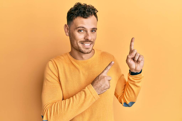 Νεαρός Ισπανός που φοράει καθημερινά ρούχα χαμογελώντας και κοιτάζοντας την κάμερα δείχνοντας με τα δύο χέρια και τα δάχτυλα στο πλάι.  - Φωτογραφία, εικόνα