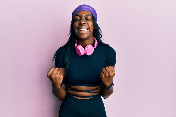 Young african american woman wearing gym clothes and using headphones very happy and excited doing winner gesture with arms raised, smiling and screaming for success. celebration concept.  - Foto, imagen
