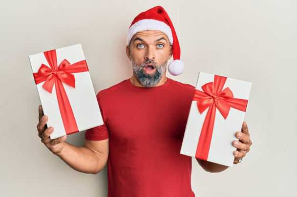 Middle age handsome man wearing christmas hat and holding a gifts in shock face, looking skeptical and sarcastic, surprised with open mouth  - Foto, Imagem
