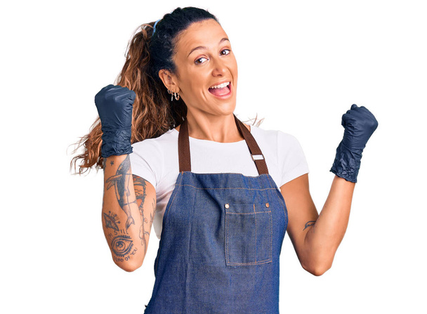 Fiatal spanyol nő tetovált borbély kötény és kesztyű visel sikoltozó büszke, ünneplő győzelem és a siker nagyon izgatott emelt karokkal  - Fotó, kép