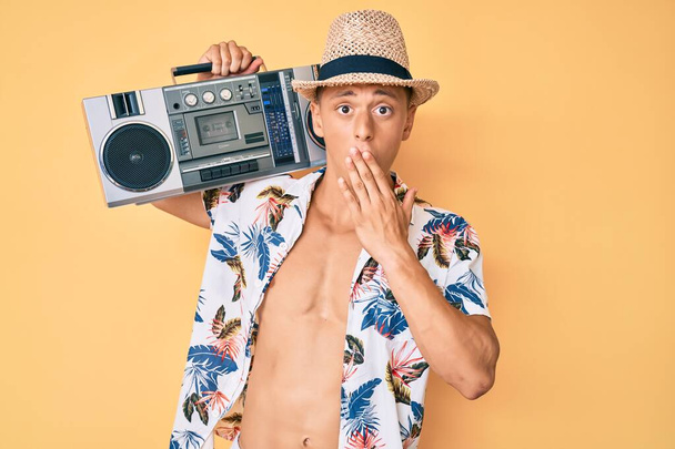 Jonge Spaanse jongen draagt zomerse stijl met boombox, luisterend naar muziek die mond met hand bedekt, geschokt en bang voor fouten. verraste uitdrukking  - Foto, afbeelding