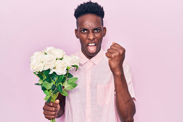 Молодий афроамериканець, який тримає квіти роздратовані і розчарований криками від гніву, з божевіллям з гнівом і піднятими руками.  - Фото, зображення
