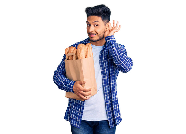 Beau jeune homme latino-américain tenant un sac en papier avec du pain souriant avec la main sur l'oreille en écoutant une rumeur ou des commérages. concept de surdité.  - Photo, image