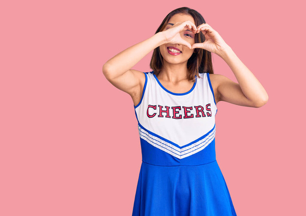 Giovane bella ragazza cinese indossa uniforme cheerleader facendo forma di cuore con la mano e le dita sorridente guardando attraverso segno  - Foto, immagini