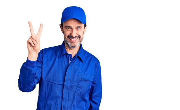 Middle age handsome man wearing mechanic uniform smiling looking to the camera showing fingers doing victory sign. number two.  - Photo, Image