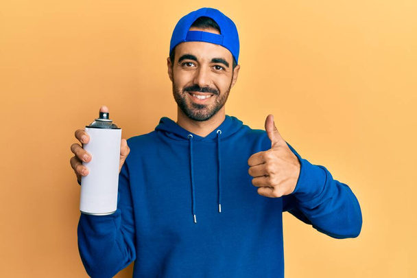 Jonge Latijns-Amerikaanse man draagt sweater met graffiti spray glimlachen blij en positief, duim omhoog doen uitstekend en goedkeuring teken  - Foto, afbeelding