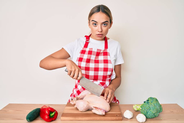 Young beautiful blonde woman wearing cook apron cutting chicken skeptic and nervous, frowning upset because of problem. negative person.  - Photo, Image