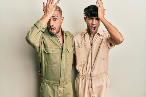 Homosexual gay couple standing together wearing casual jumpsuit surprised with hand on head for mistake, remember error. forgot, bad memory concept.  - Foto, Bild