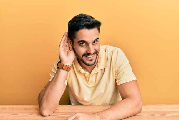 Young hispanic man wearing casual clothes sitting on the table smiling with hand over ear listening an hearing to rumor or gossip. deafness concept.  - Photo, Image