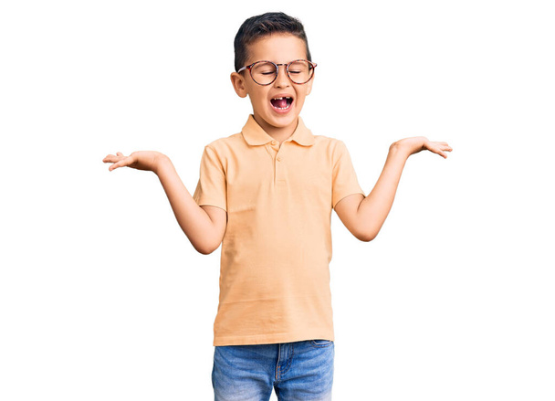 Μικρό χαριτωμένο αγόρι παιδί φορώντας casual ρούχα και γυαλιά γιορτάζει τη νίκη με χαρούμενο χαμόγελο και την έκφραση του νικητή με υψωμένα χέρια  - Φωτογραφία, εικόνα
