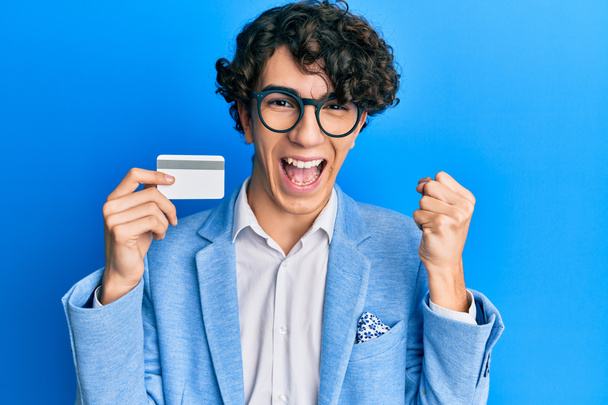 Hispanic young man holding credit card screaming proud, celebrating victory and success very excited with raised arms  - Foto, Imagen