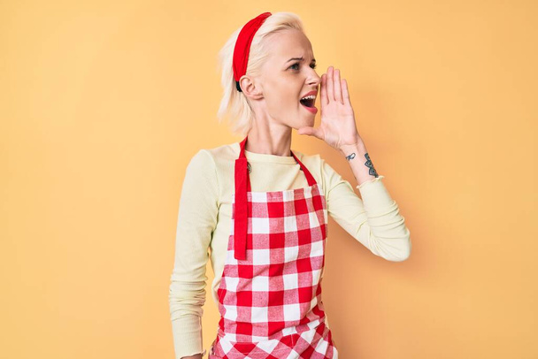 Jeune femme blonde avec tatouage portant tablier boulanger professionnel criant et criant fort à côté avec la main sur la bouche. concept de communication.  - Photo, image