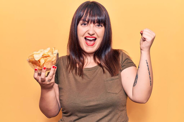 Junge Plus-Size-Frau mit Kartoffelchips in der Hand schreit stolz und feiert Sieg und Erfolg sehr aufgeregt mit erhobenen Armen  - Foto, Bild