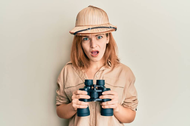 Jeune femme caucasienne portant un chapeau explorateur regardant à travers les jumelles dans le visage de choc, regardant sceptique et sarcastique, surpris avec la bouche ouverte  - Photo, image