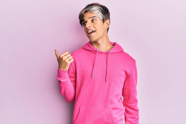Joven hombre hispano con sudadera rosa casual apuntando con el pulgar hacia un lado sonriendo feliz con la boca abierta  - Foto, imagen