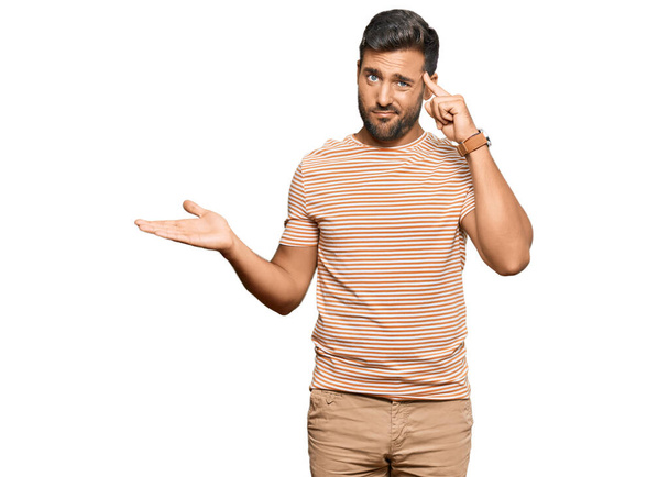 Hombre hispano guapo vistiendo ropa casual confundido y molesto con la palma abierta mostrando espacio de copia y señalando el dedo a la frente. Piénsalo.  - Foto, Imagen