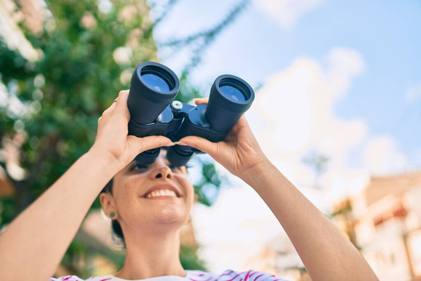 Young hispanic girl smiling happy looking for new opportunity using binoculars at the city. - 写真・画像